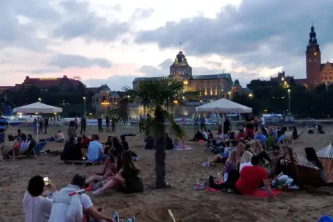 Ludzie odpoczywają na plaży, ciesząc się wieczorem. W tle widać piękne budynki, świadczące o bogactwie wydarzeń kulturalnych w Szczecinie.